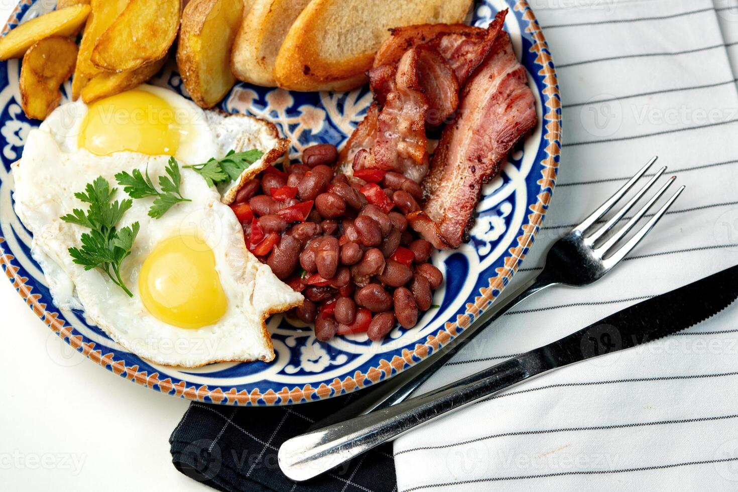 Delicious breakfast plate featuring eggs, beans, bacon, and potato wedges served on a decorative dish photo