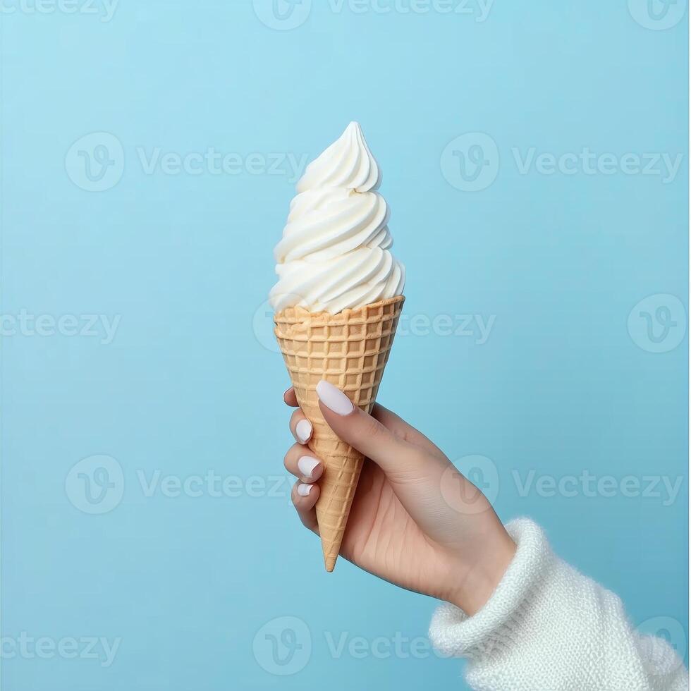 A hand holding a swirl of soft serve ice cream in a cone against a light blue backdrop during a sunny day photo
