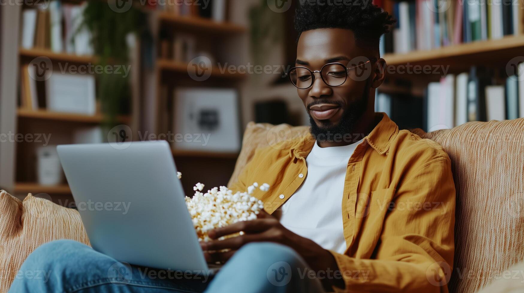 A man enjoys a cozy evening at home, browsing on a laptop while relaxing with a bowl of popcorn in a well-lit living room photo