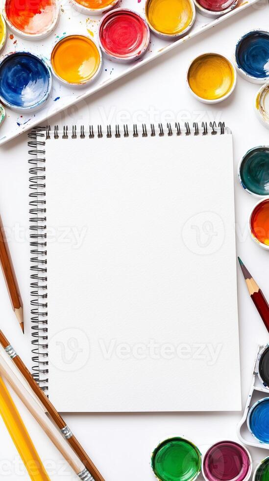 Blank sketchbook surrounded by vibrant watercolor paints and brushes on a white table photo
