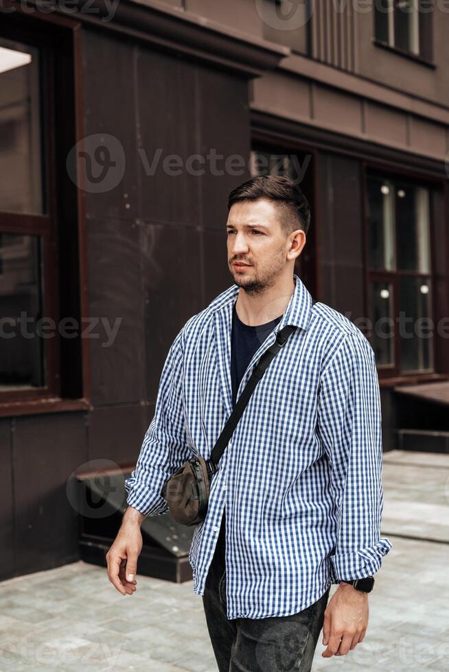 A man in a blue checkered shirt smiles while standing outside a modern building on a sunny day photo