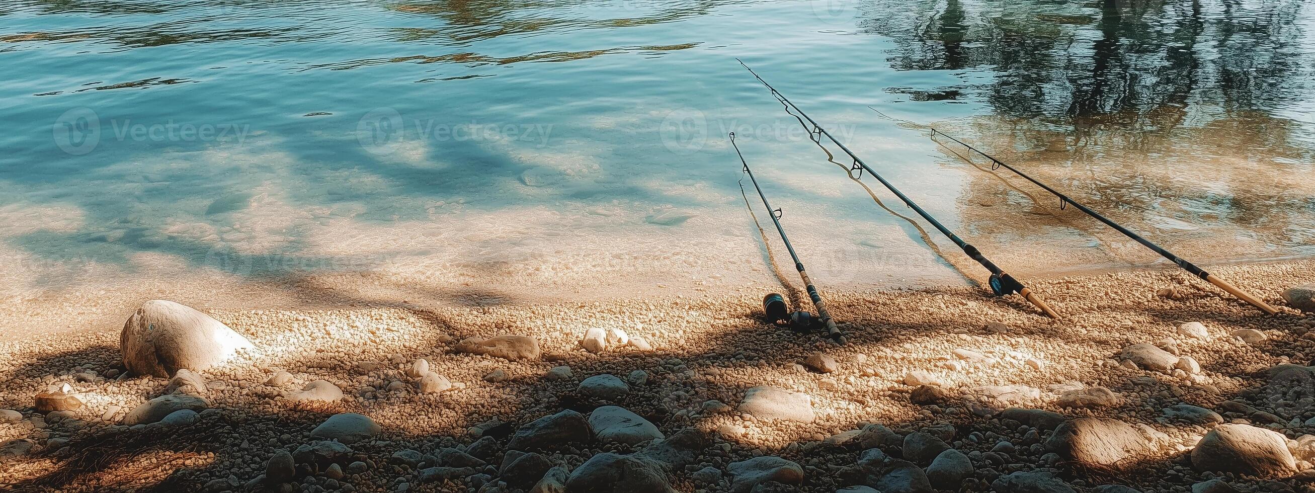 Tranquil riverbank in early morning light with fishing rods resting on sandy shore surrounded by reflections and gentle water ripples photo