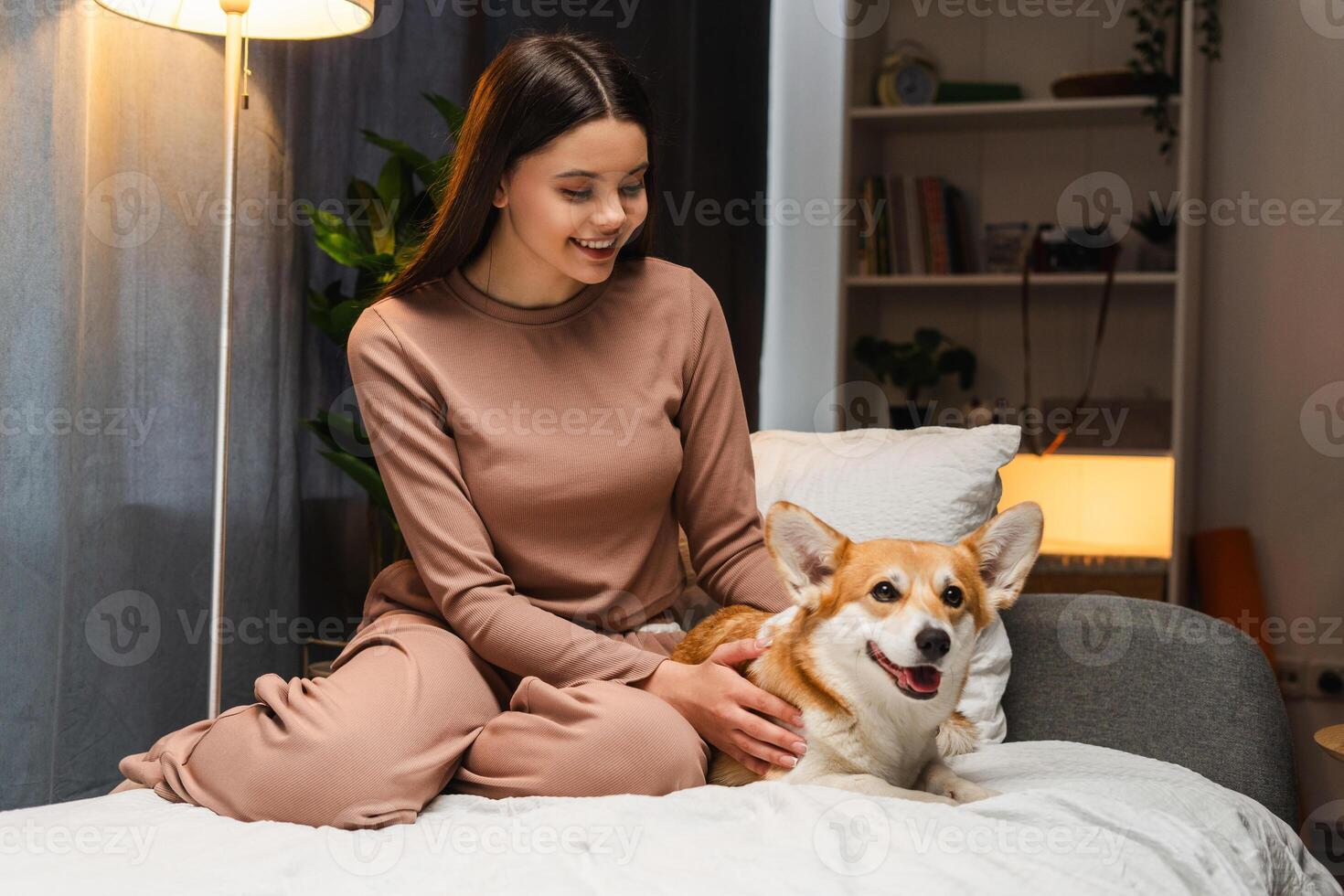 Smiling young girl, teenagerpetting her corgi dog on bed at home photo
