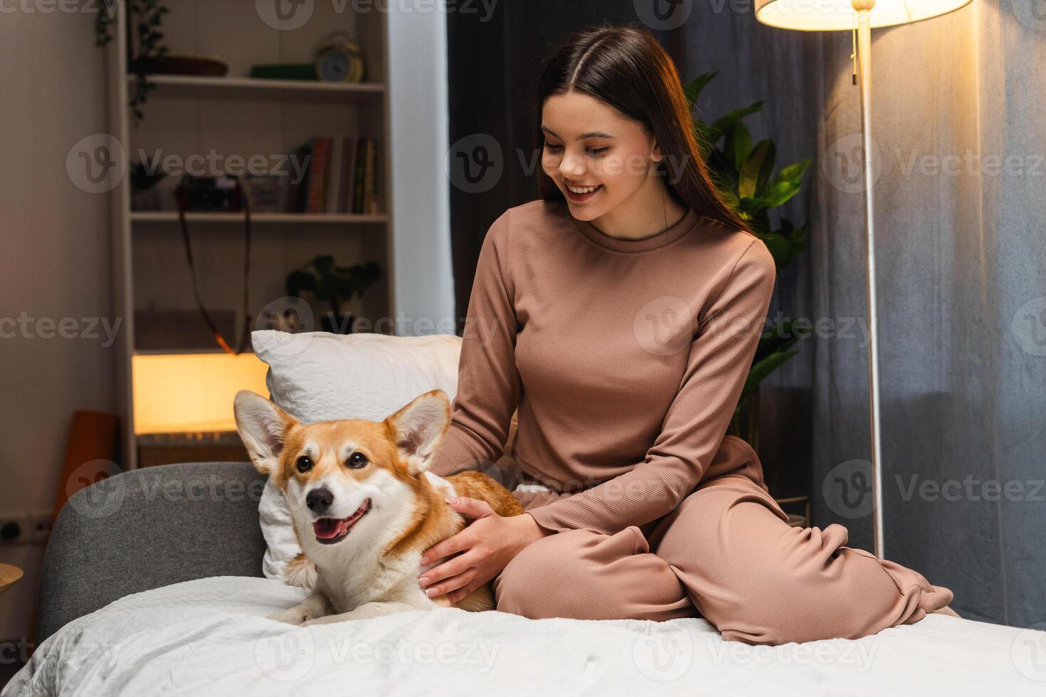 Smiling young girl, teenagerpetting her corgi dog on bed at home photo
