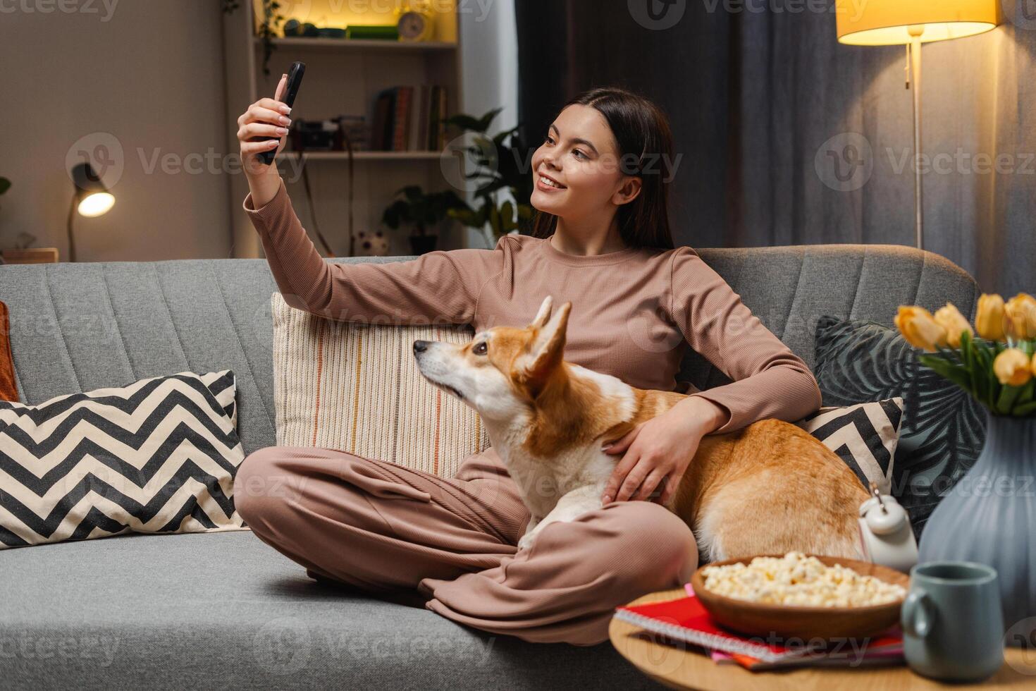 Smiling girl relaxing on sofa with corgi and taking selfie with smartphone photo