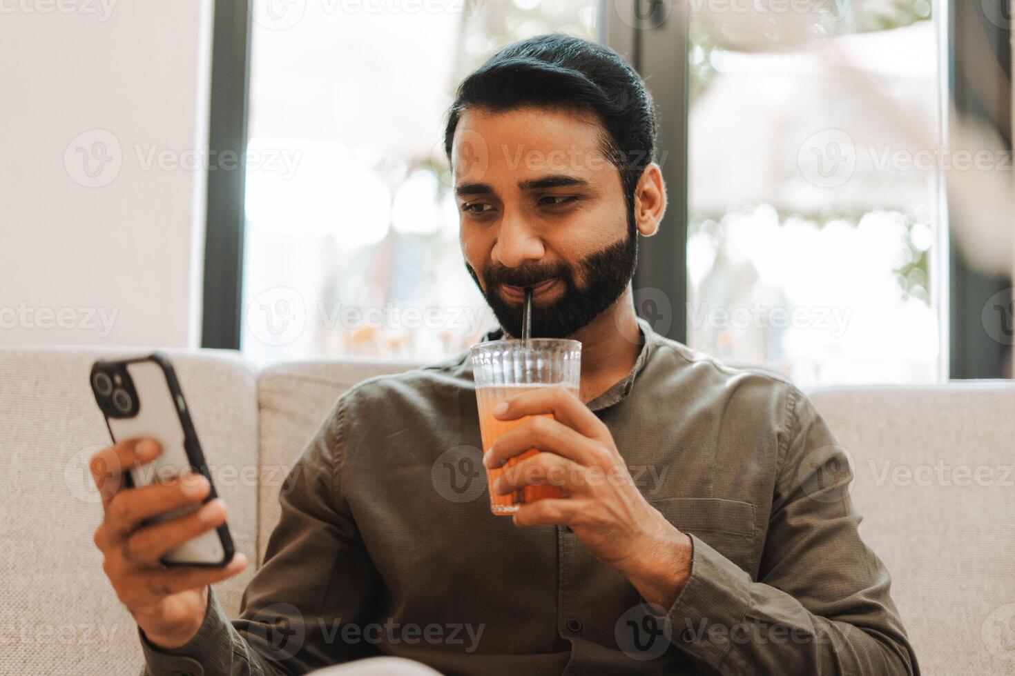 Happy, handsome Indian man using mobile phone and drinking juice on sofa in cafe photo