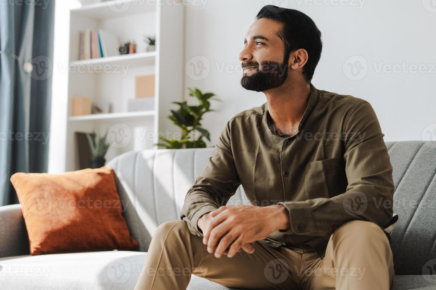 Handsome attractive Indian businessman sitting on sofa at home looking away and smiling photo