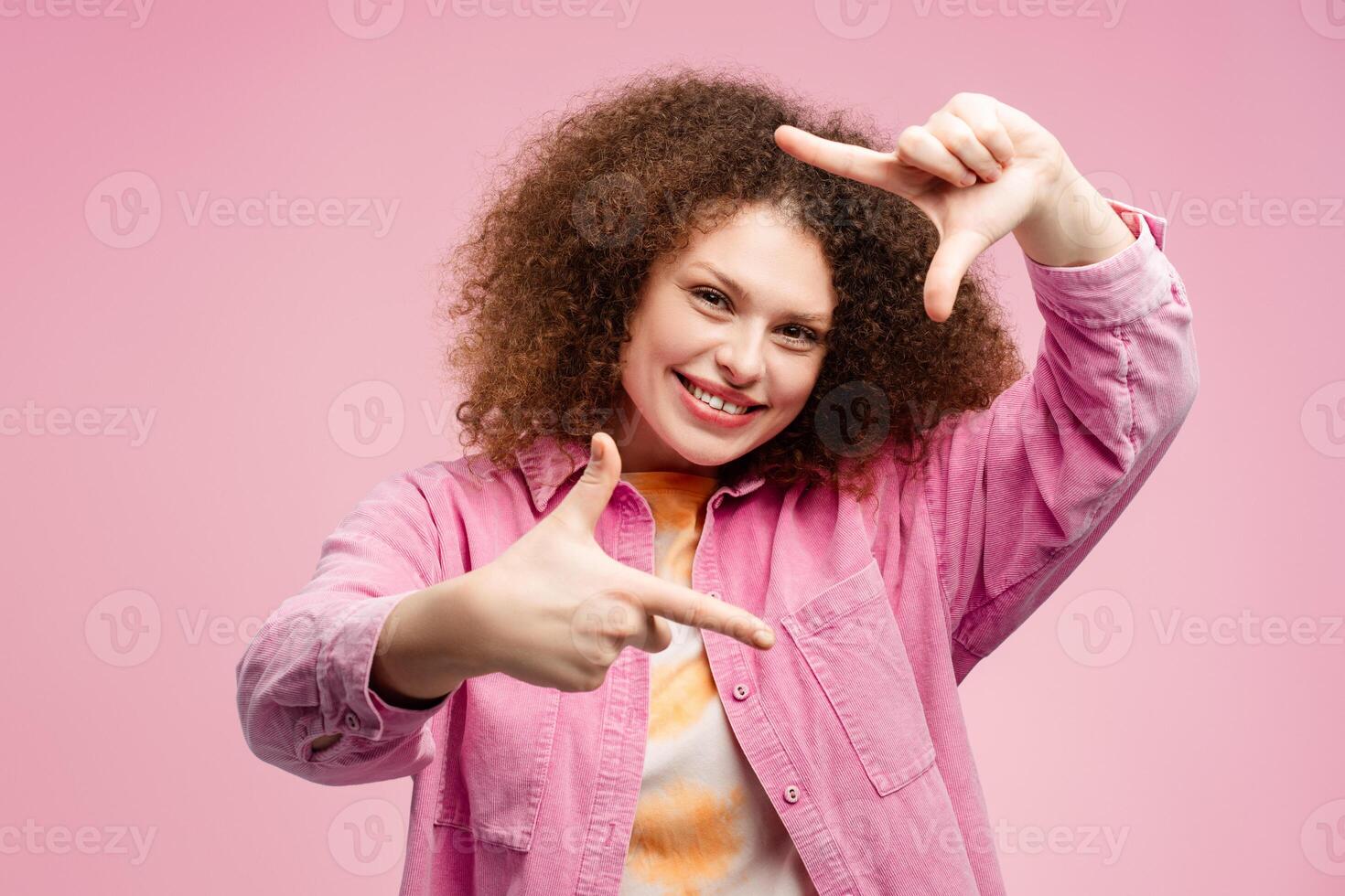 Smiling, young woman with curly hair shows frame with her fingers, gesturing looking at camera photo