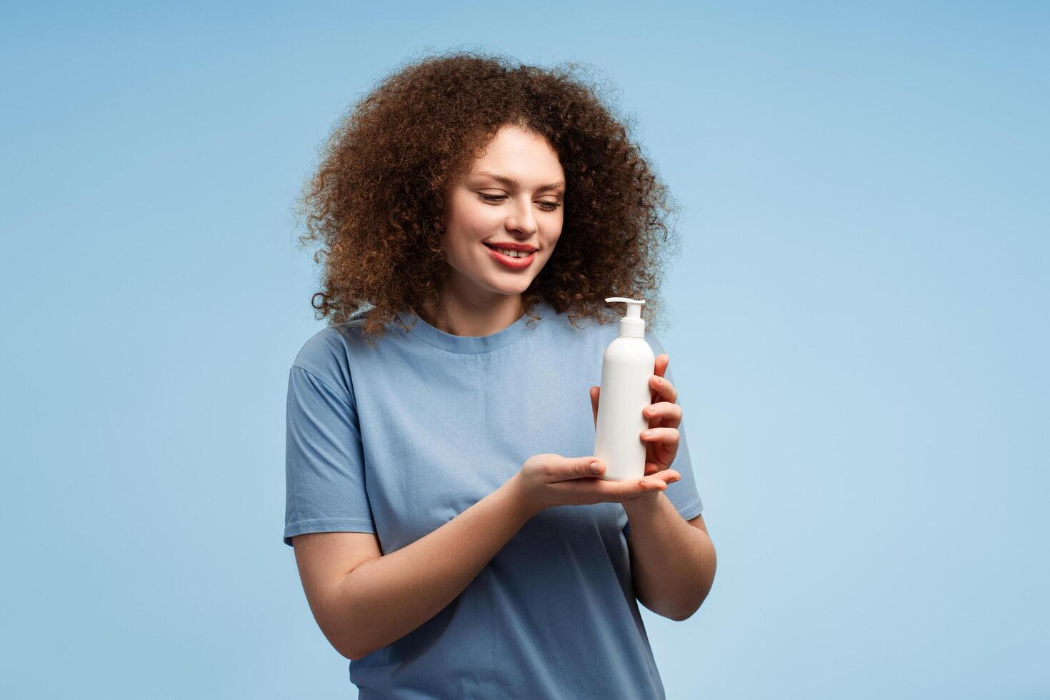 Smiling, happy woman with curly hair holding white shampoo bottle, mockup photo