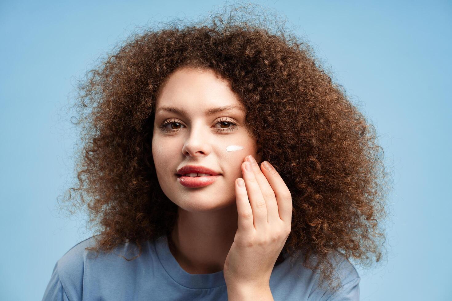 Smiling beautiful young woman with curly hair applying cream on face, looking in mirror photo