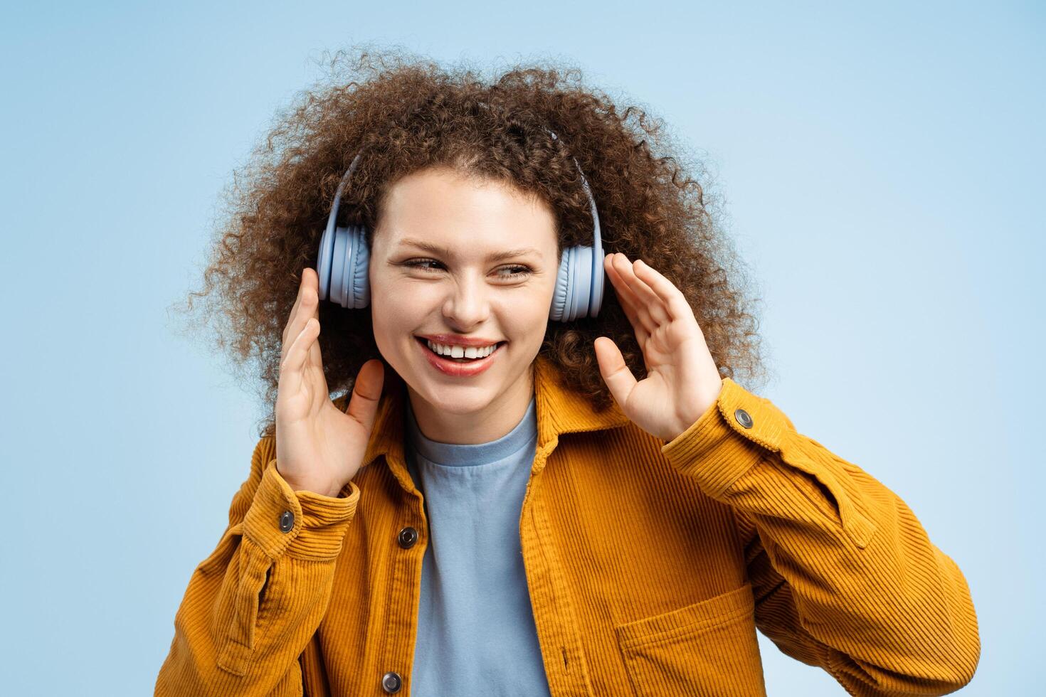 Young woman with curly hair wearing stylish yellow jacket listening to music in wireless headphones photo