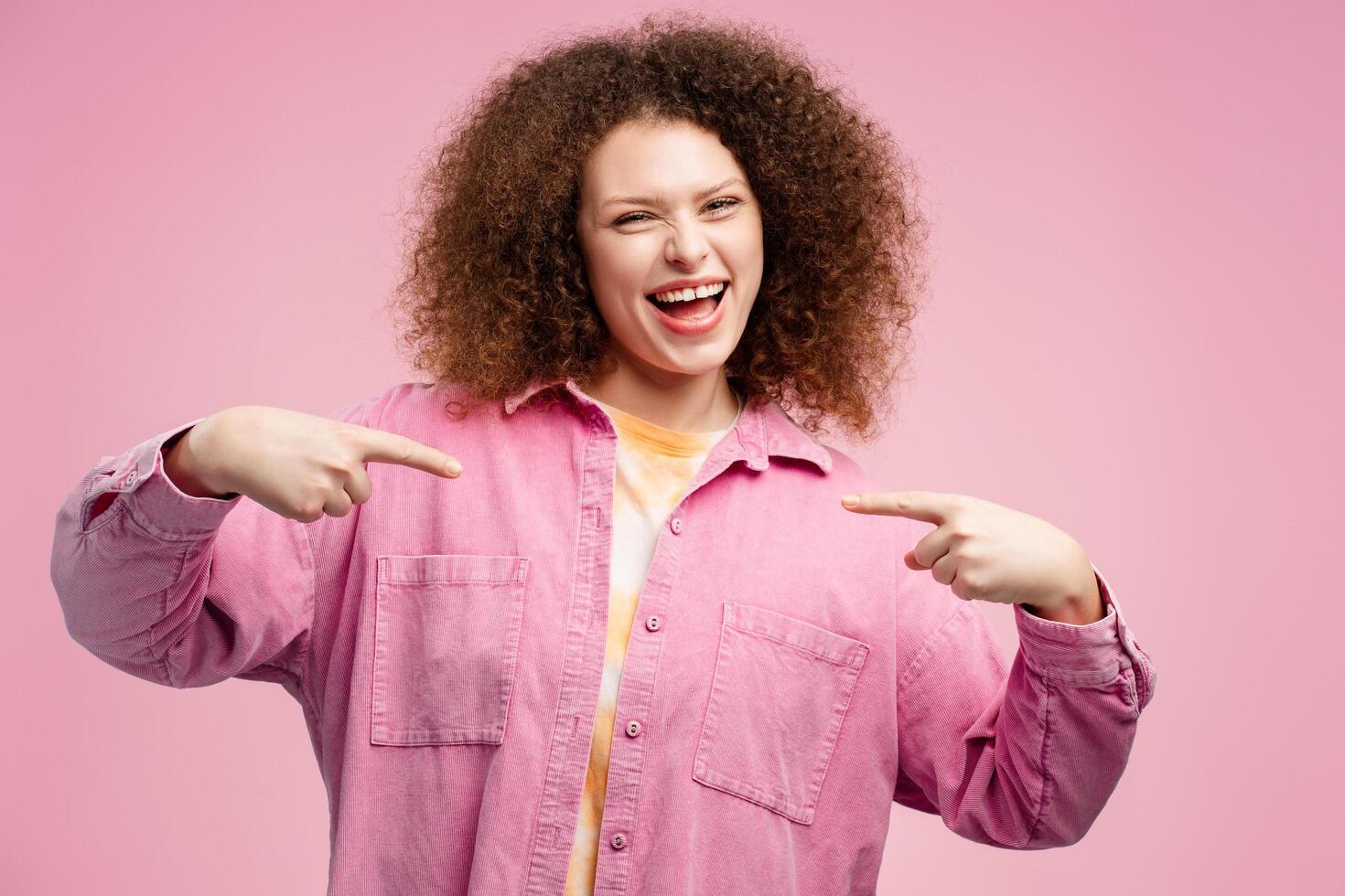 Cheerful smiling woman with curly hair pointing fingers at herself while looking at camera photo