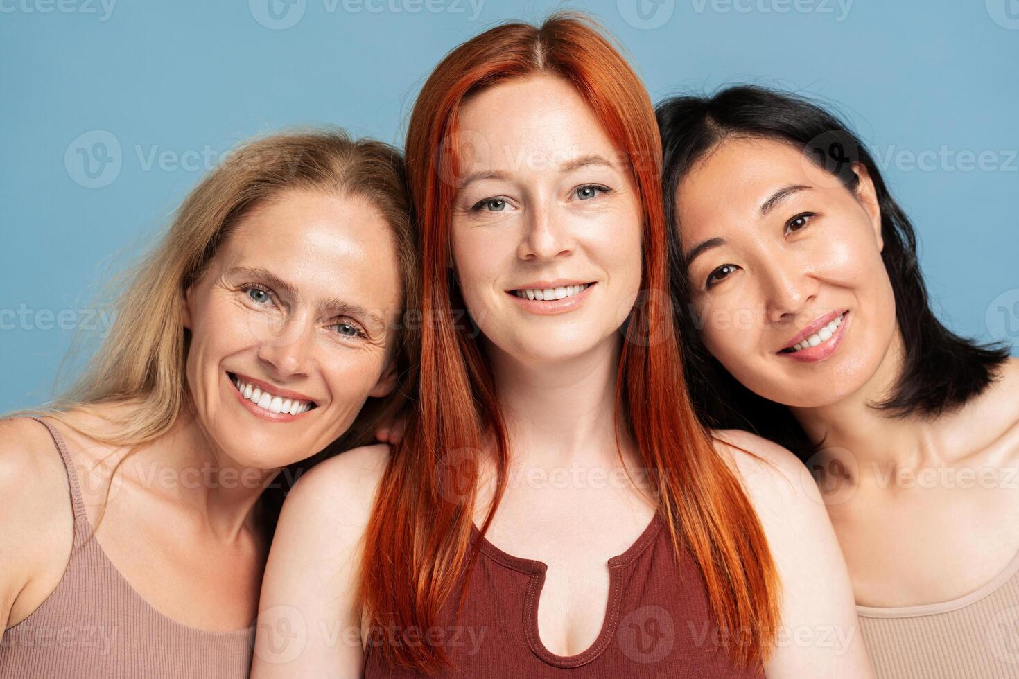Three confident women smiling and embracing looking at camera, posing isolated on blue background photo