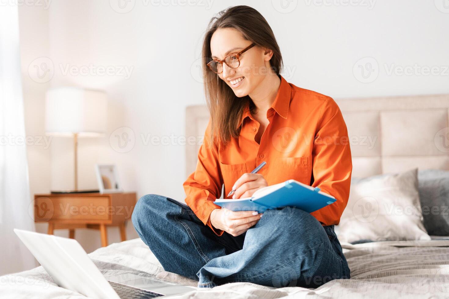 Student woman in eyeglasses sitting on bed using laptop, writing in notebook, studying photo