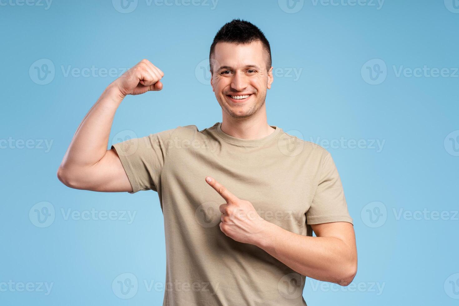 Strong, athletic, smiling man in pastel t shirt pointing biceps, showing strength photo