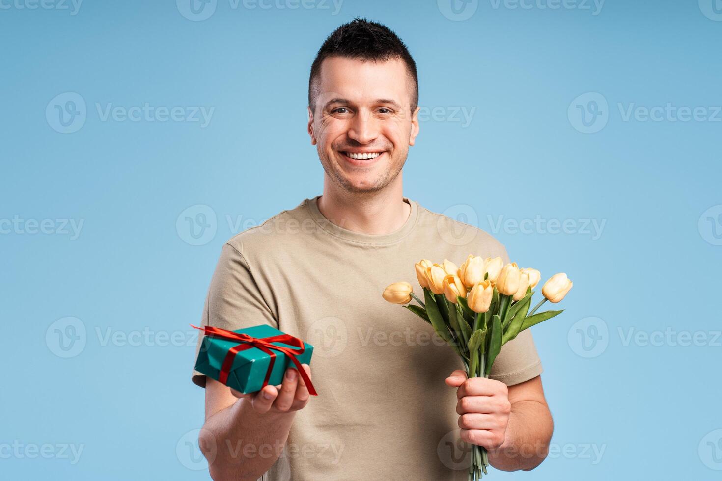 Handsome smiling young man looking at camera, holding bouquet of flowers and gift in hand photo
