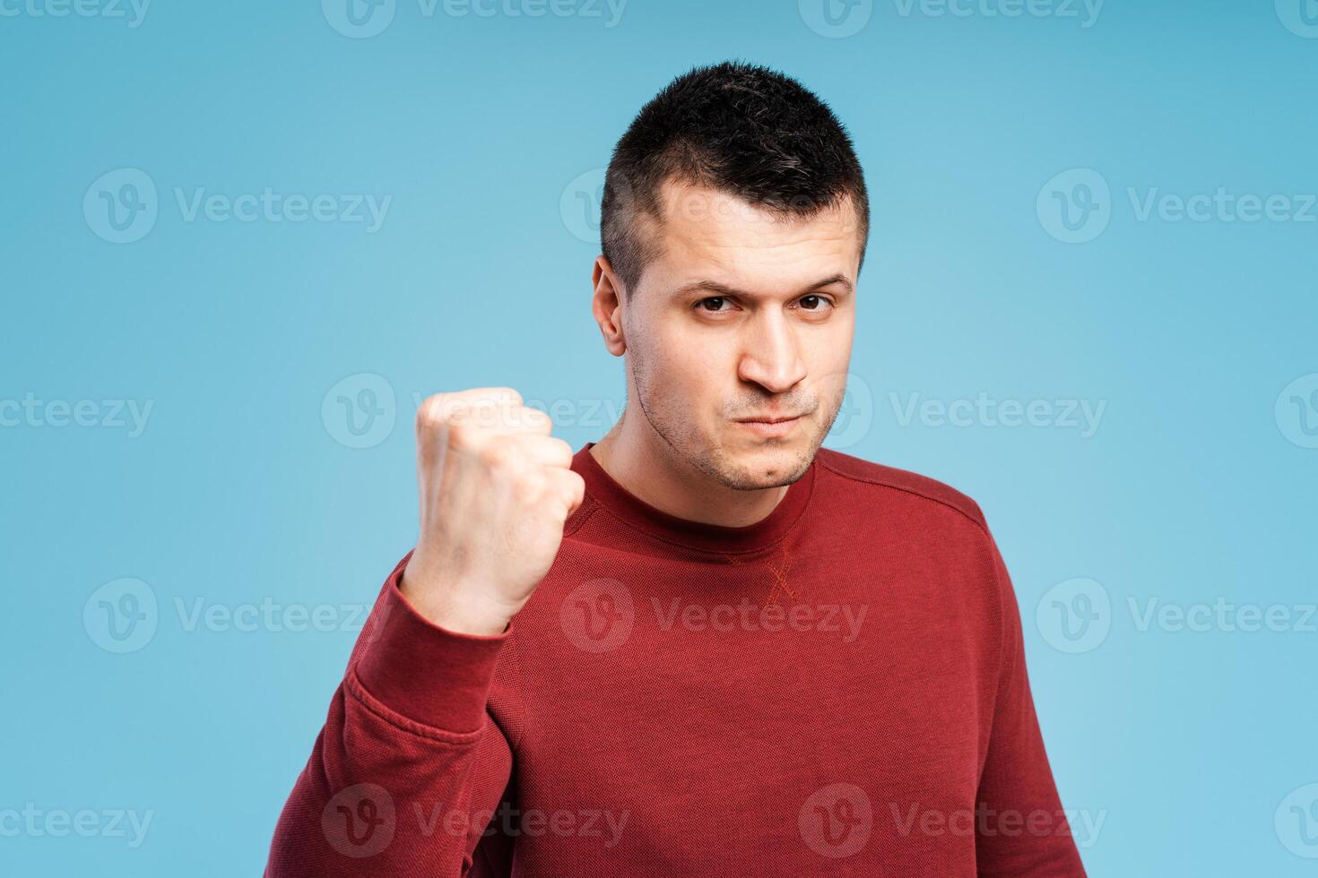 Angry, irritated young man in red sweatshirt showing clenched fist, showing anger. Conflict concept photo