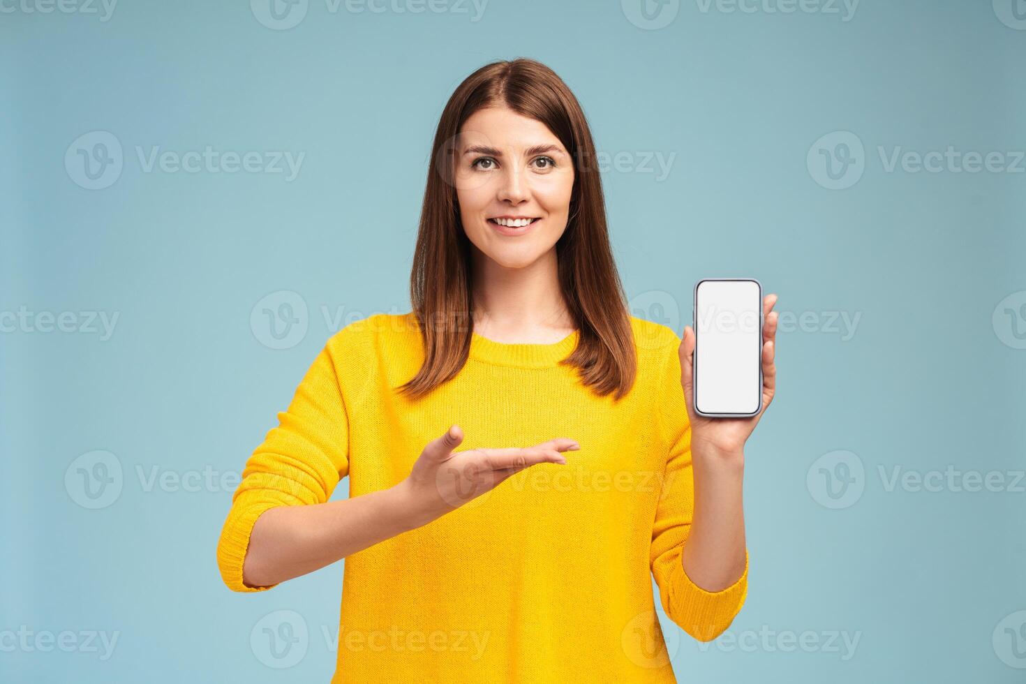 Beautiful smiling Caucasian woman holding mobile phone, pointing to white blank screen, mockup photo