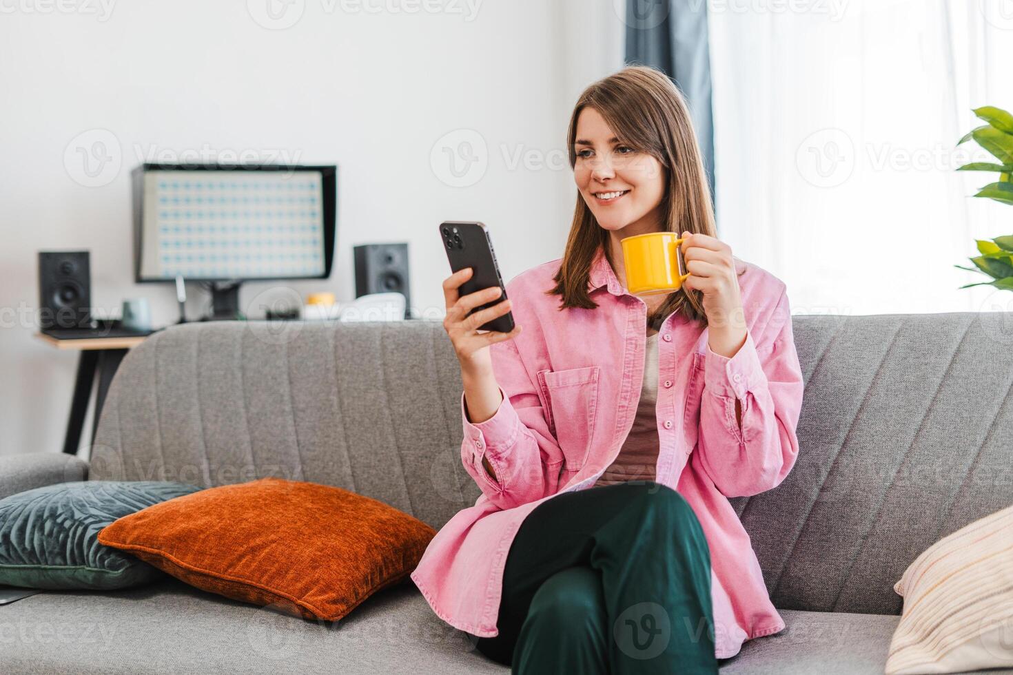 Portrait of attractive, smiling woman sitting on bed at home holding mobile phone, using mobile app photo