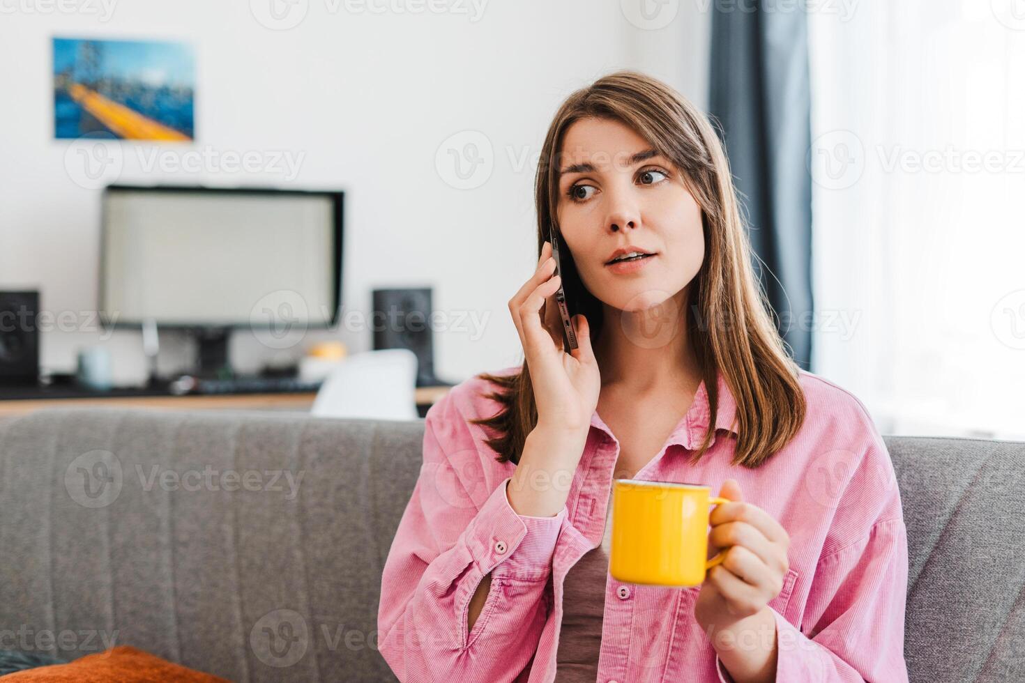 Smiling woman answering call, using mobile phone and yellow cup, sitting on comfortable sofa at home photo