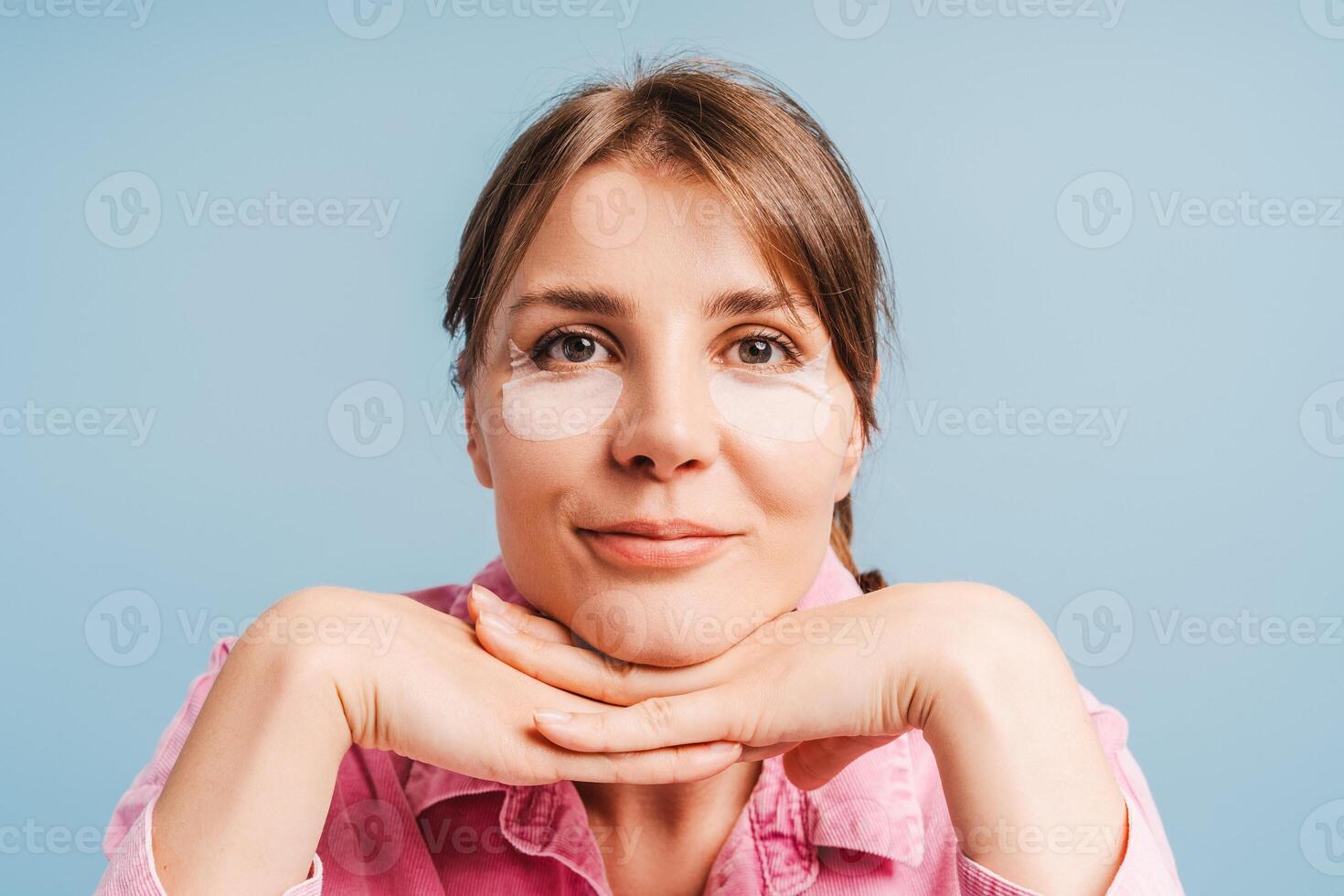 Smiling woman with patches under her eyes looking in mirror standing isolated on blue background photo