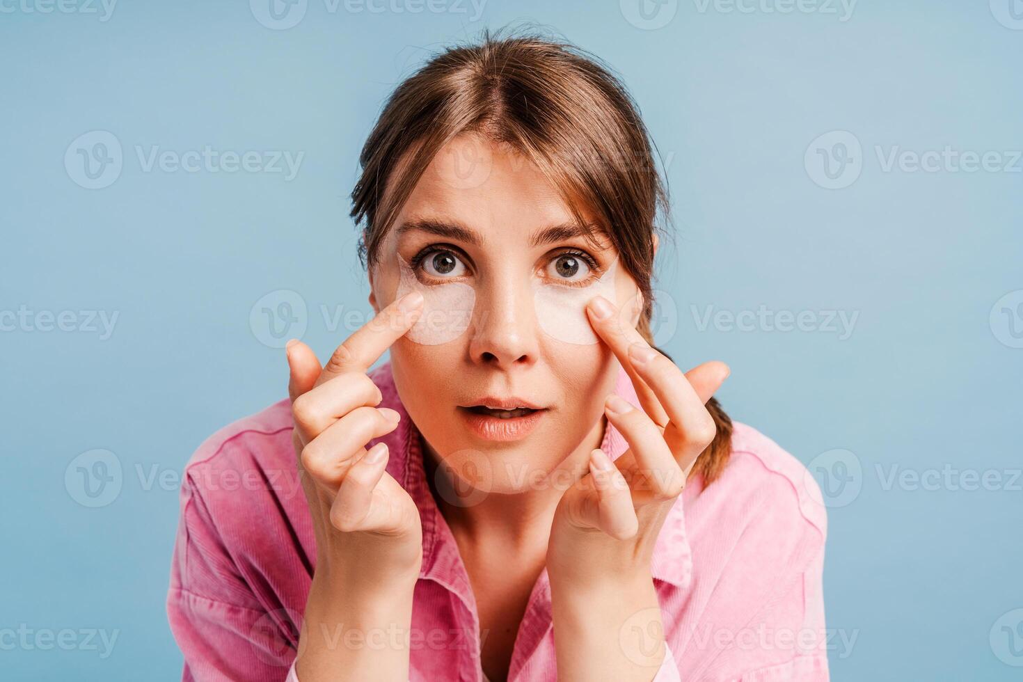 Beautiful woman with patches under eyes looking in mirror, posing isolated on blue background photo