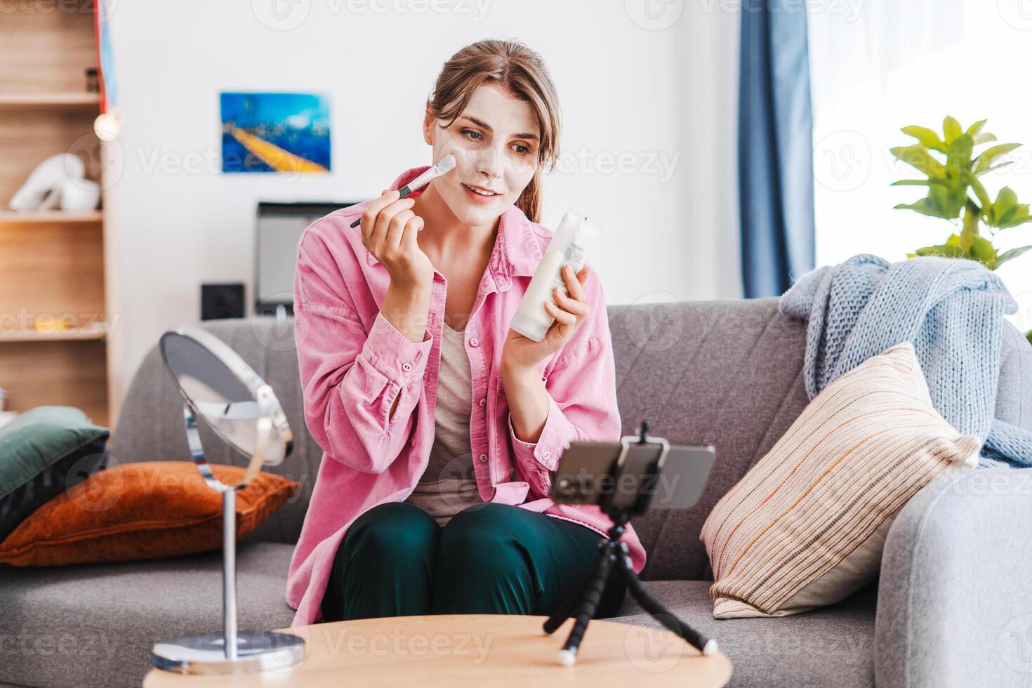 Cheerful cute woman blogger applying mask on face with brush, recording , using mobile phone photo