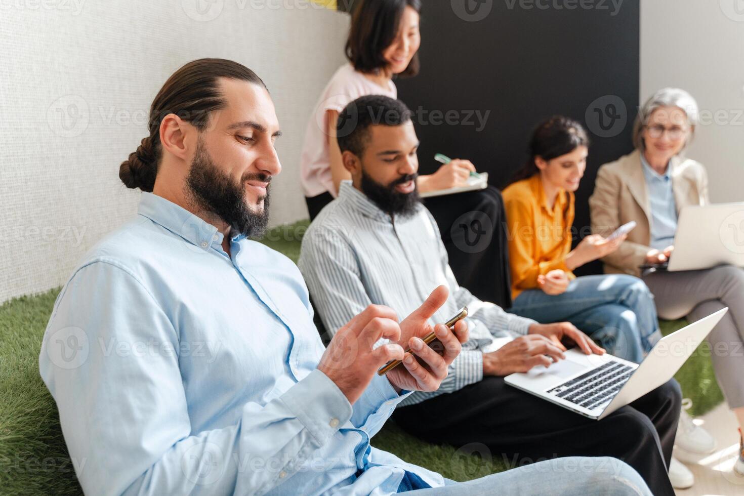 Young man using smartphone while sitting with multiethnic coworkers on sofa photo
