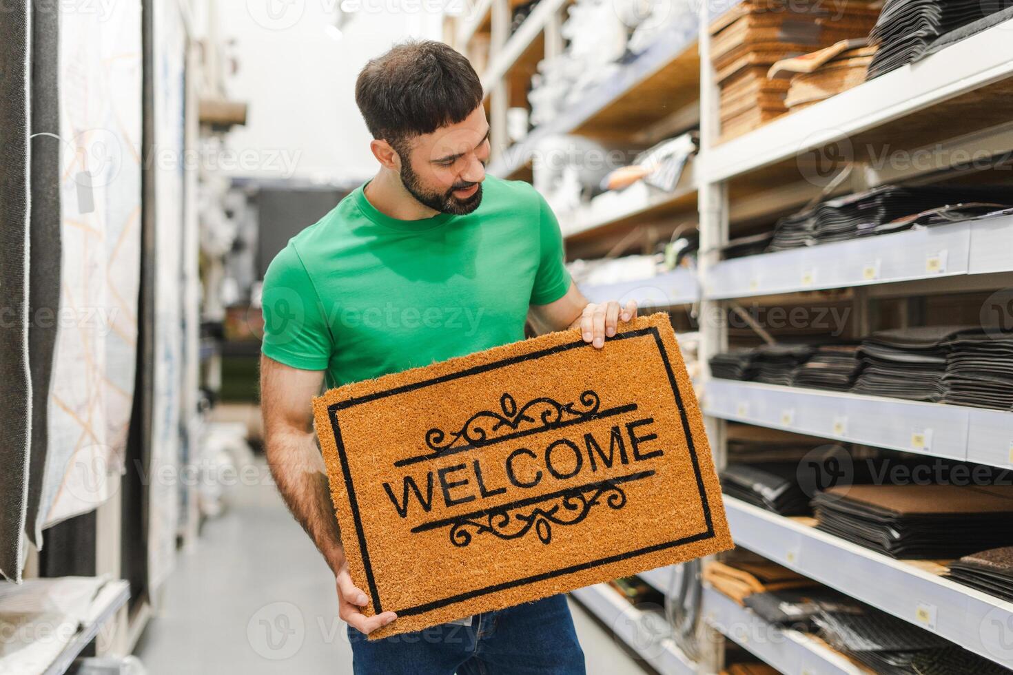 Happy young bearded man picking new door mat with word Welcome in decor department photo