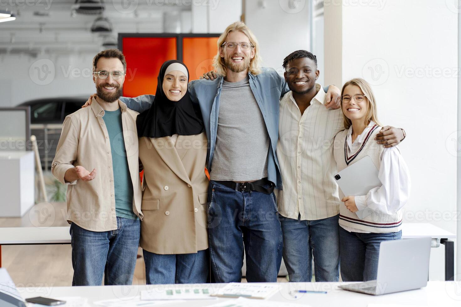 Diverse multiracial team of five business people hugging posing in the office, looking at camera photo