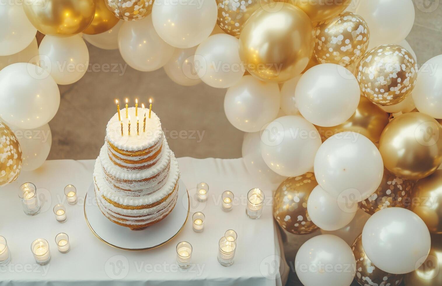 An elegant birthday party background featuring gold and white balloons, a table with a tiered cake, and candles adding a soft glow photo