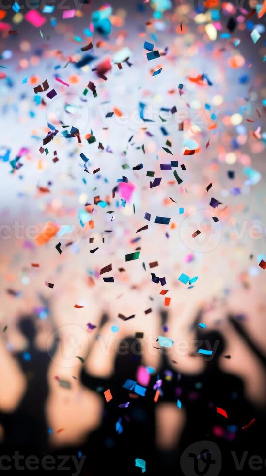 A close-up of falling confetti in focus, with blurred silhouettes of people celebrating in the background photo