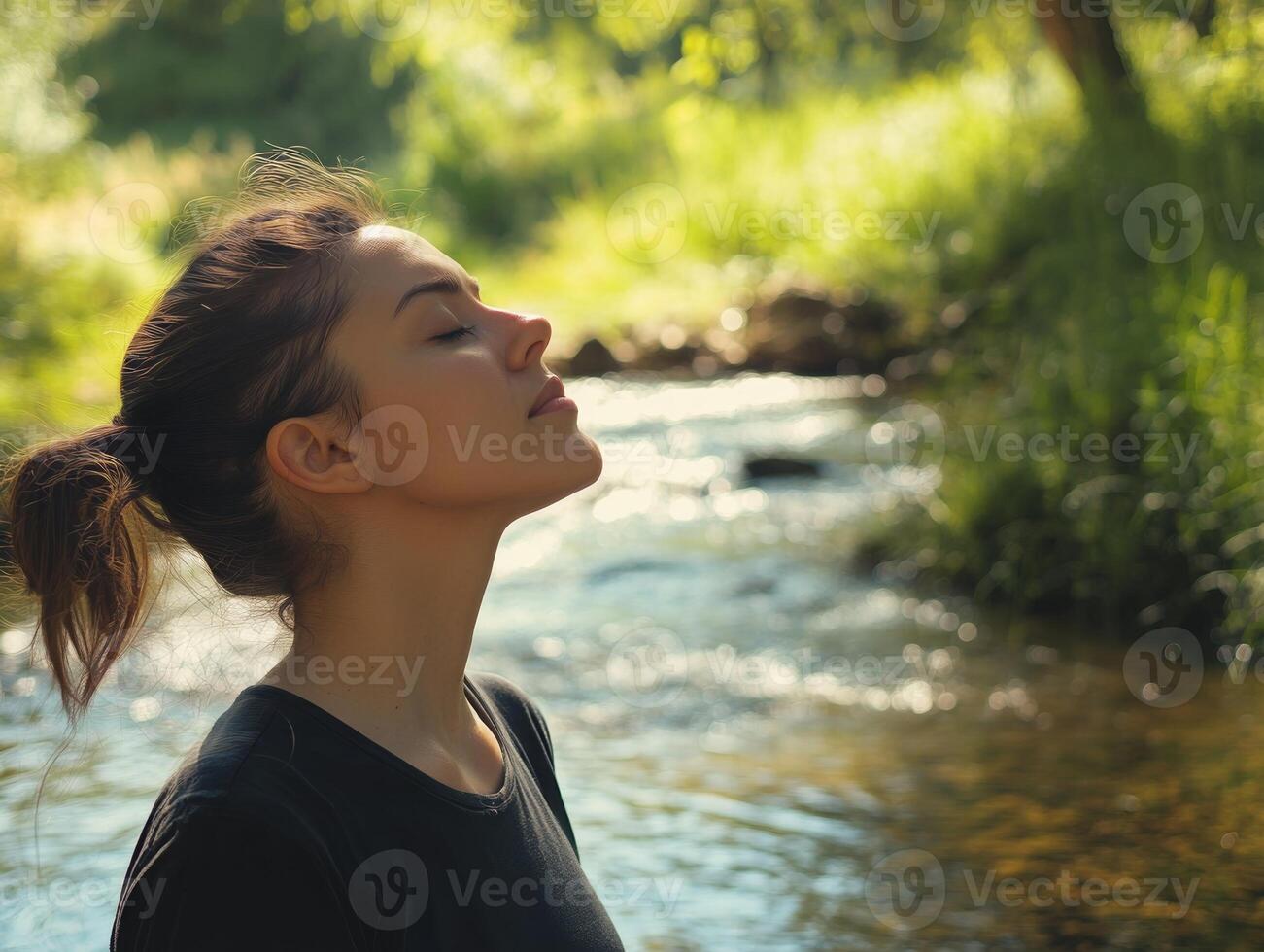 Woman outdoors, standing by a stream with her eyes closed, head tilted back, breathing deeply, with space for copy in the peaceful landscape photo