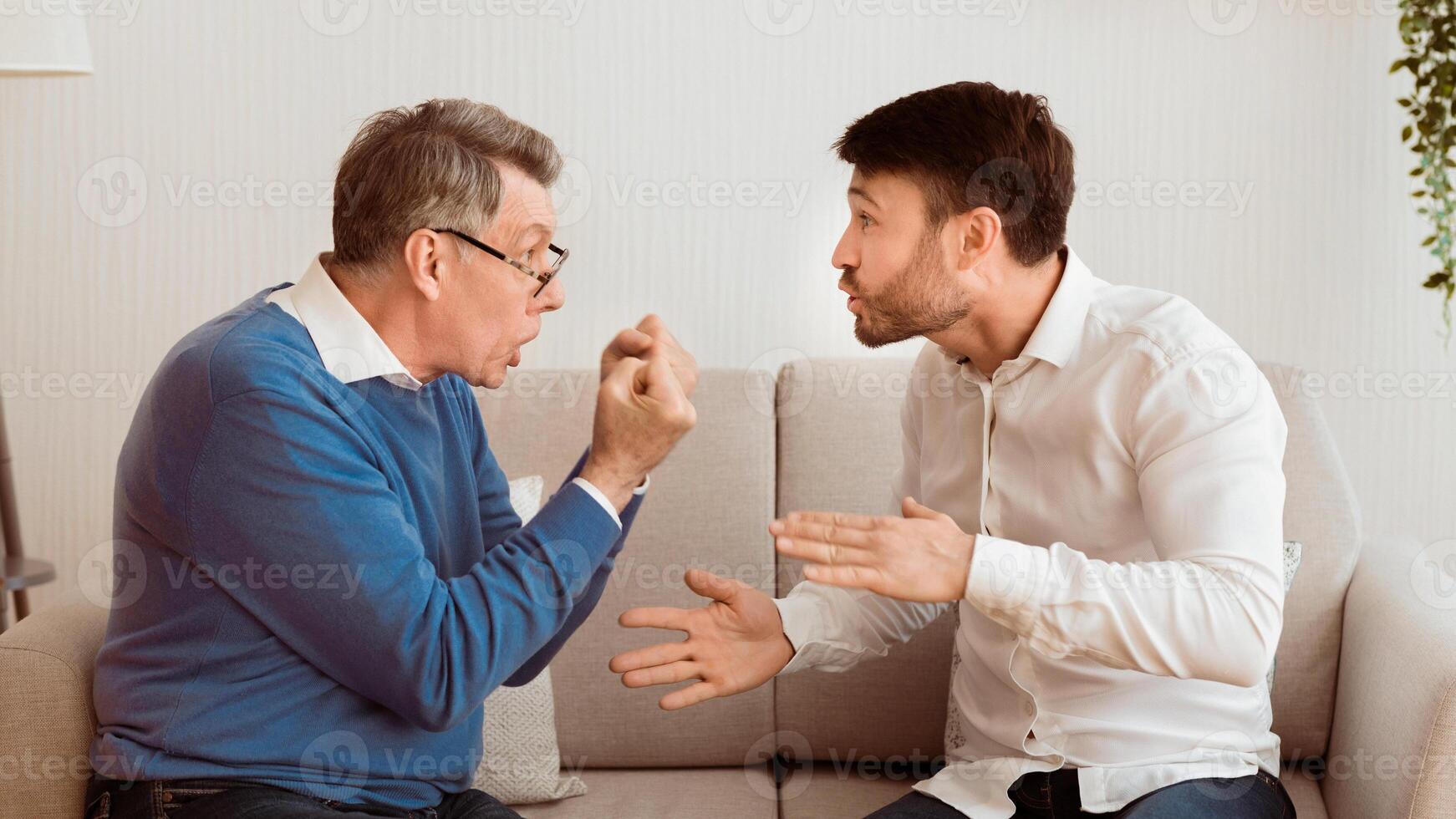 Father And Son Quarrel. Aggressive Senior Man Having Argument With Adult Son Sitting On Couch Indoor. Panorama, Selective Focus photo