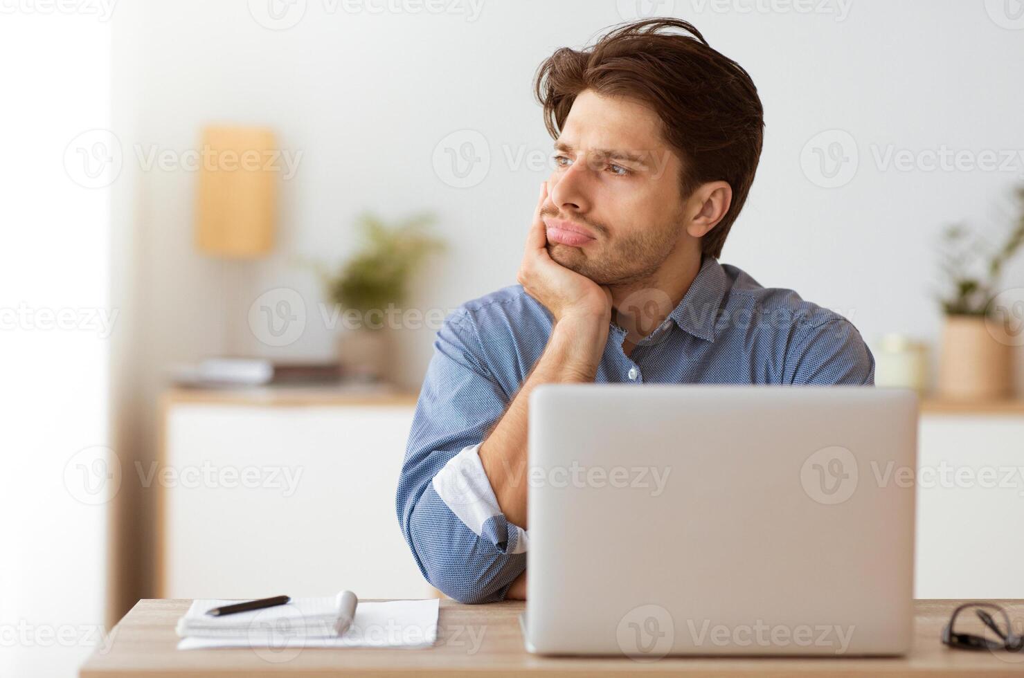 A man sits at a desk with his hand on his chin, looking thoughtfully away from the laptop in front of him. photo