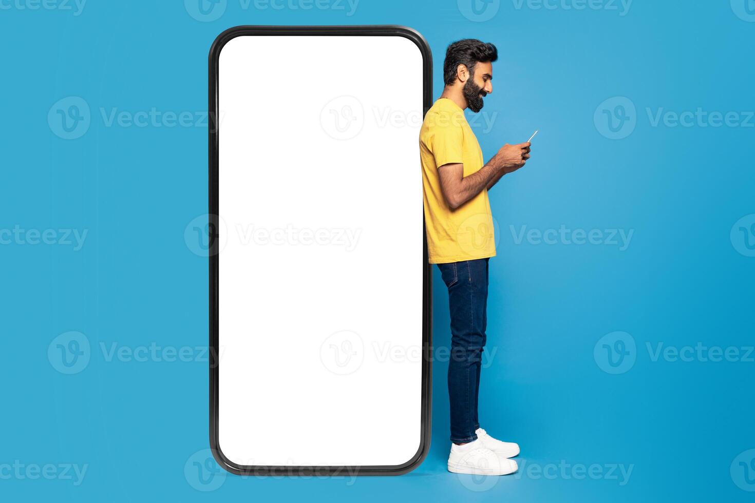 A young Indian man wearing a yellow shirt and jeans is engrossed in using his smartphone. He is standing against a giant phone frame with a blank white screen, set against a vivid blue background photo
