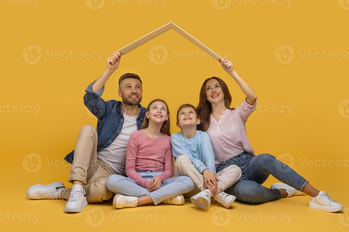A smiling family sits closely together with the parents holding their arms up to form the shape of a house over their two children heads, all set against a vibrant yellow backdrop photo