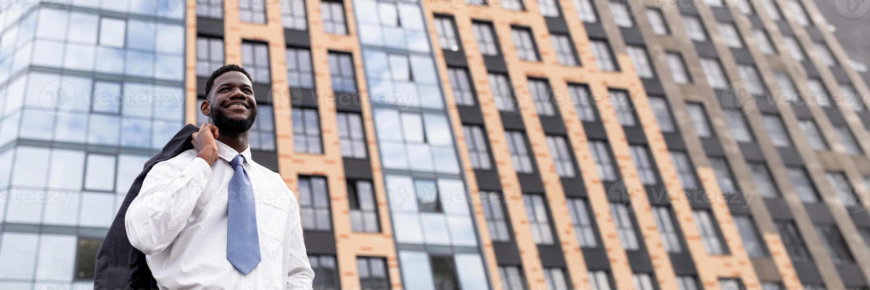 Happy african american businessman holding jacket on shoulder and leaving modern office building, free space, web-banner photo