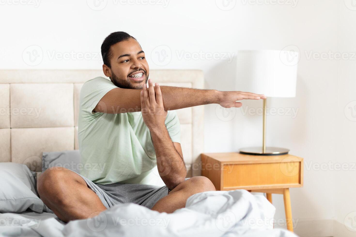 A young African American man is sitting on a bed, wearing casual sleepwear and stretching with a content smile photo