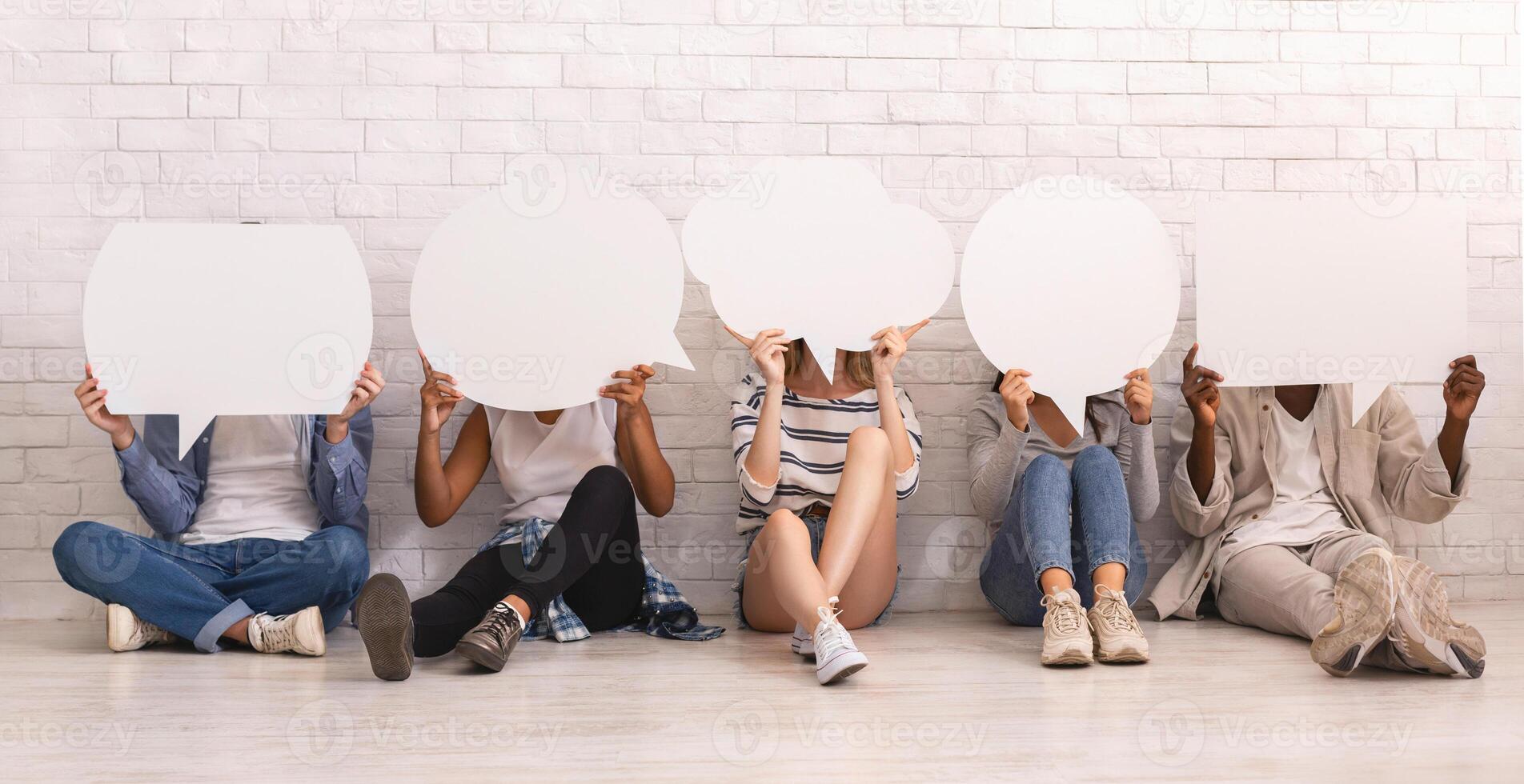 Group of young people sitting on floor over white brick wall, covering faces with speech bubbles, panorama photo