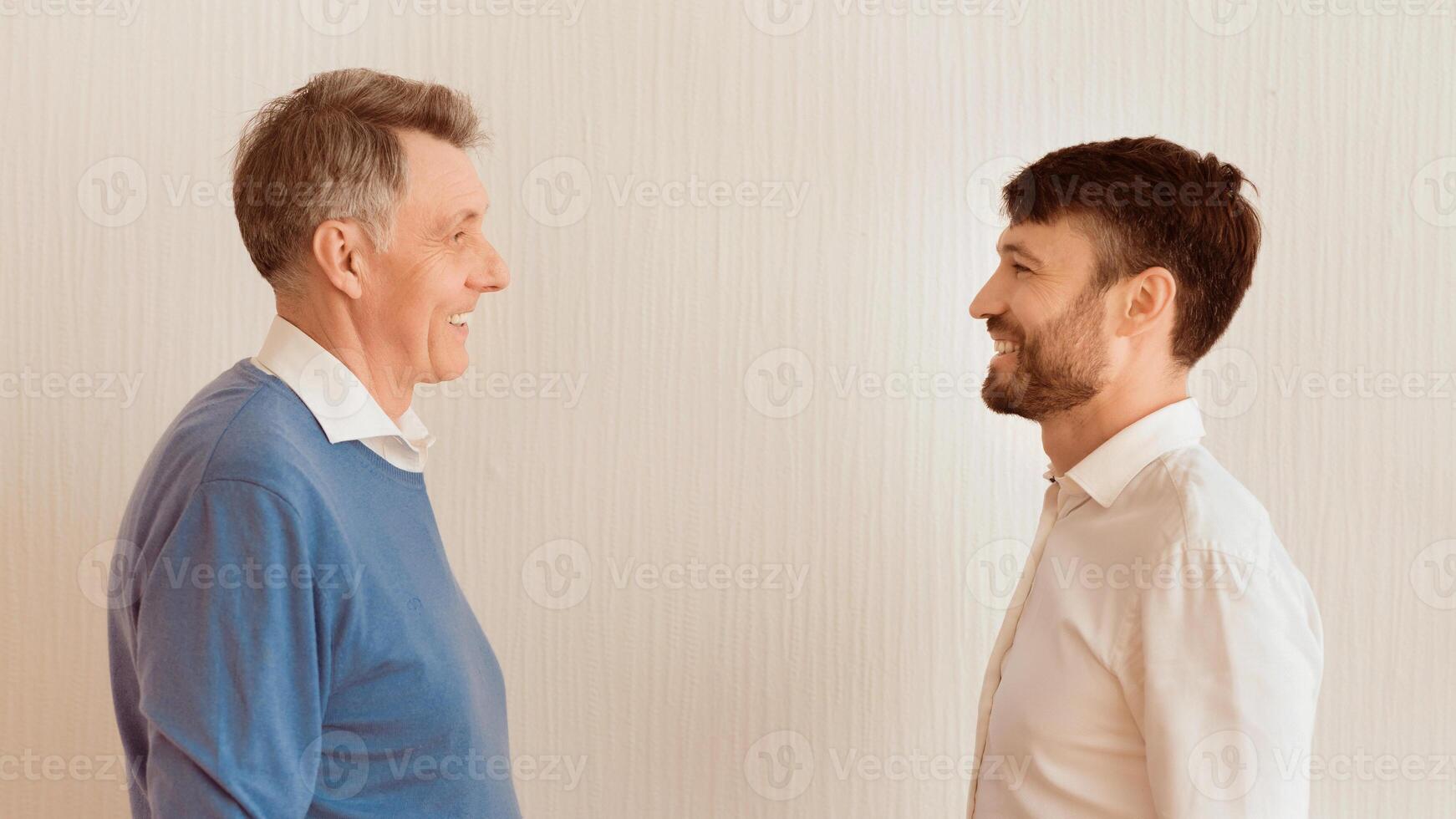 Mature Man And His Elderly Dad Smiling Each Other Standing Against White Wall Indoor. Father And Adult Son Relationship. Panorama photo