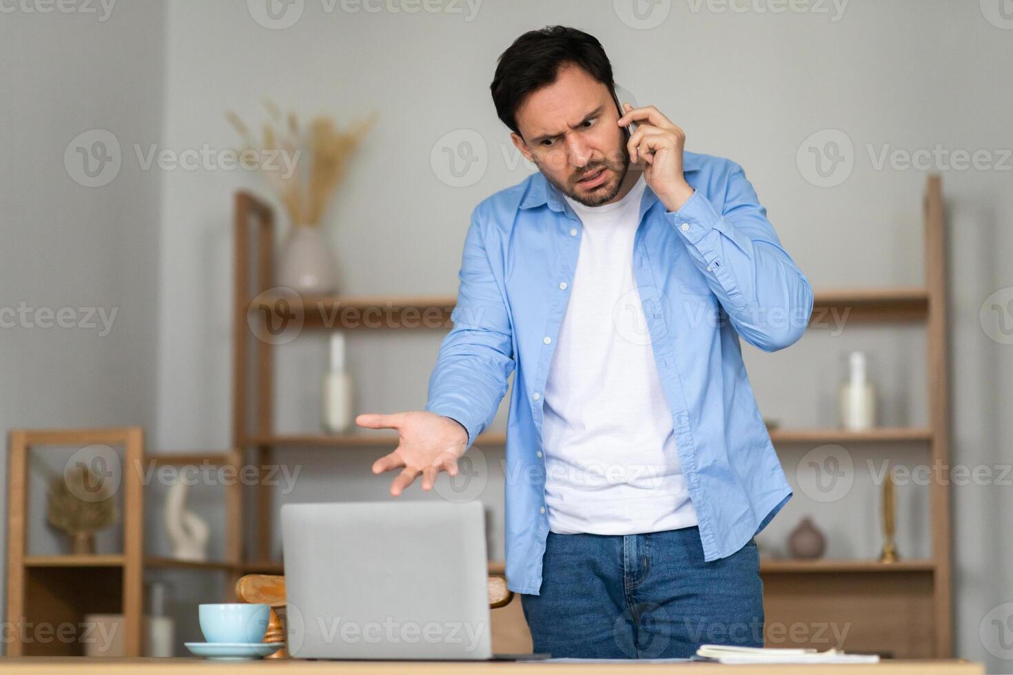 A man stands in front of his laptop in a home office, expressing frustration and confusion during a phone call. His body language, with one hand on his hip and a furrowed brow photo