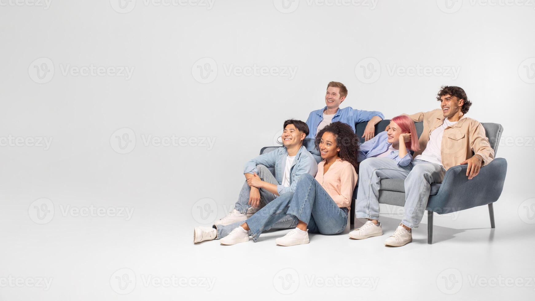 A group of five diverse friends are relaxing and enjoying each others company in a studio setting. They are sitting on a grey couch and the floor, laughing and looking happy, copy space photo