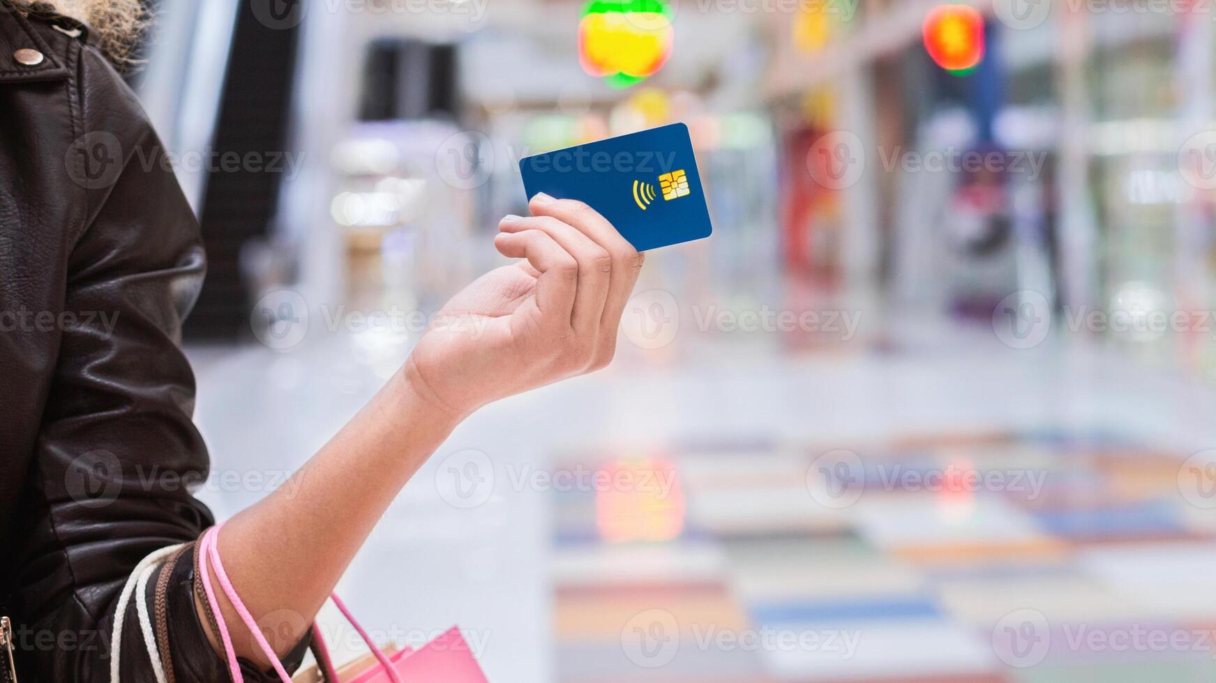 A woman is standing in a busy shopping mall, holding a credit card in one hand. She appears focused on making a purchase or payment, surrounded by shops and other shoppers, cropped photo