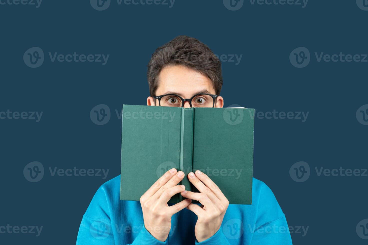 A young man sits against a solid dark blue background, holding an open book in front of his face. He wears glasses and a bright blue sweater, peering over the top of the book photo