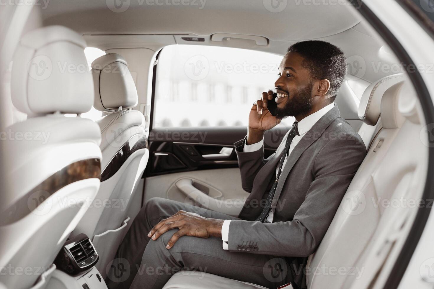 African American businessman in a suit is sitting in the backseat of a car, talking on his phone with a smile on his face. He is dressed in a gray suit, white shirt, and a dark tie photo