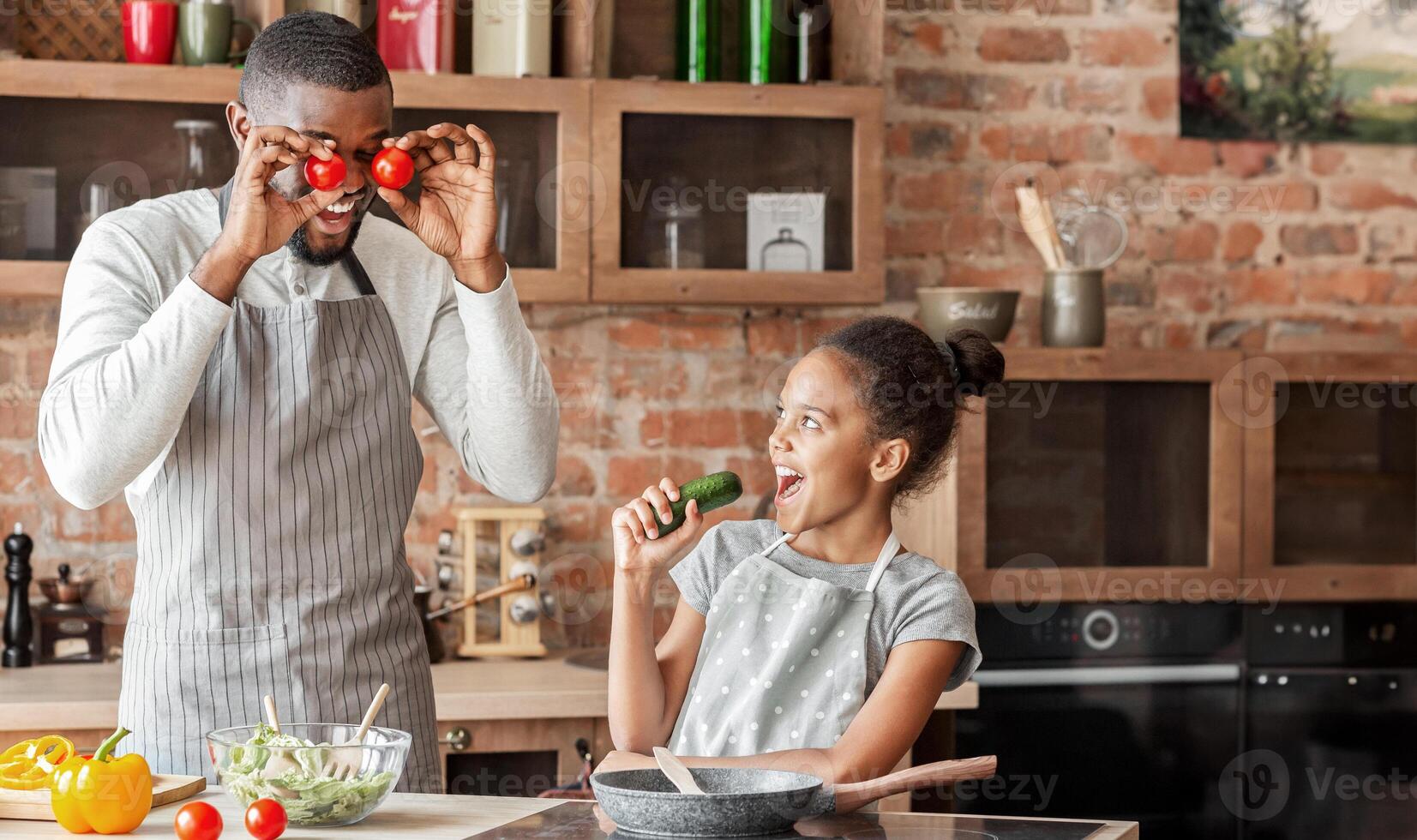 Afro man and his daughter having fun at kitchen, girl singing in cucumber, dad making tomato eyes, empty space photo