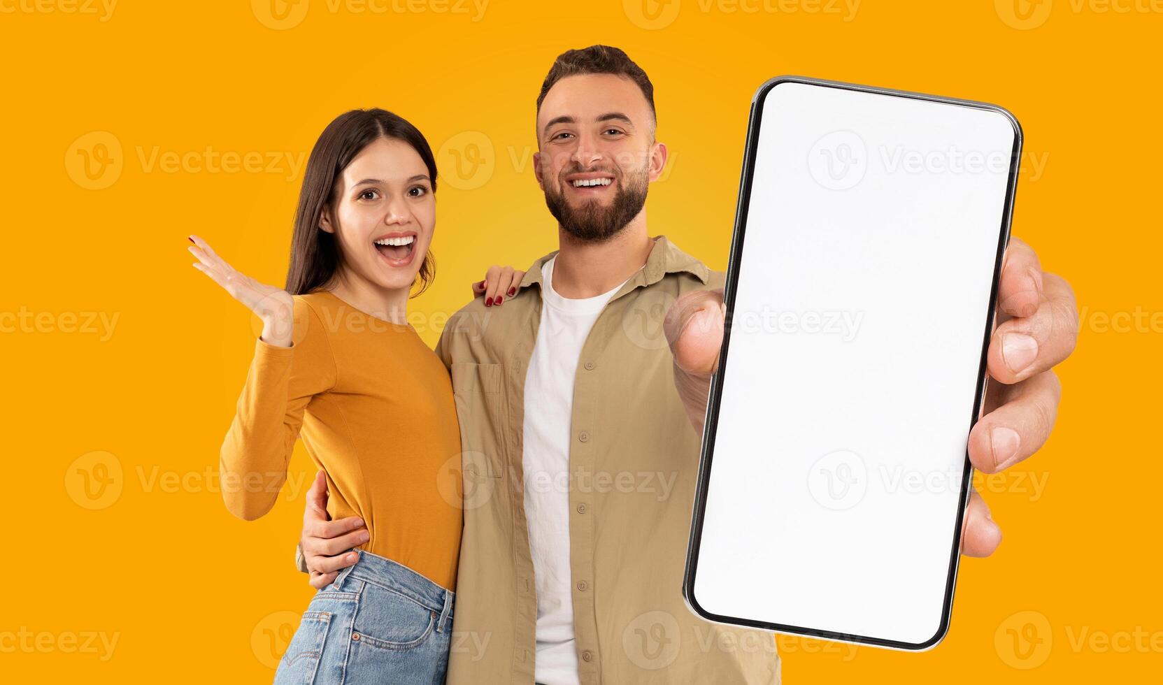 A young couple standing in front of a bright yellow background. The man is holding a smartphone with a blank screen in front of him, and the woman is standing beside him photo