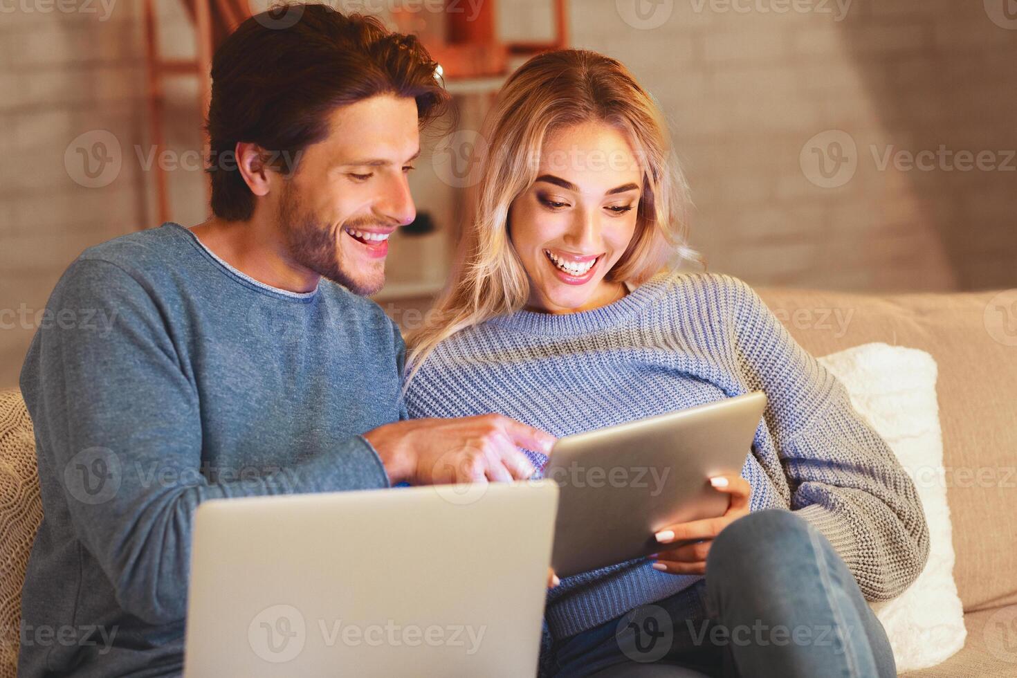 Smiling Man And Woman Using Laptop And Digital Tablet Websurfing And Chatting In Social Media Sitting On Sofa Indoor photo