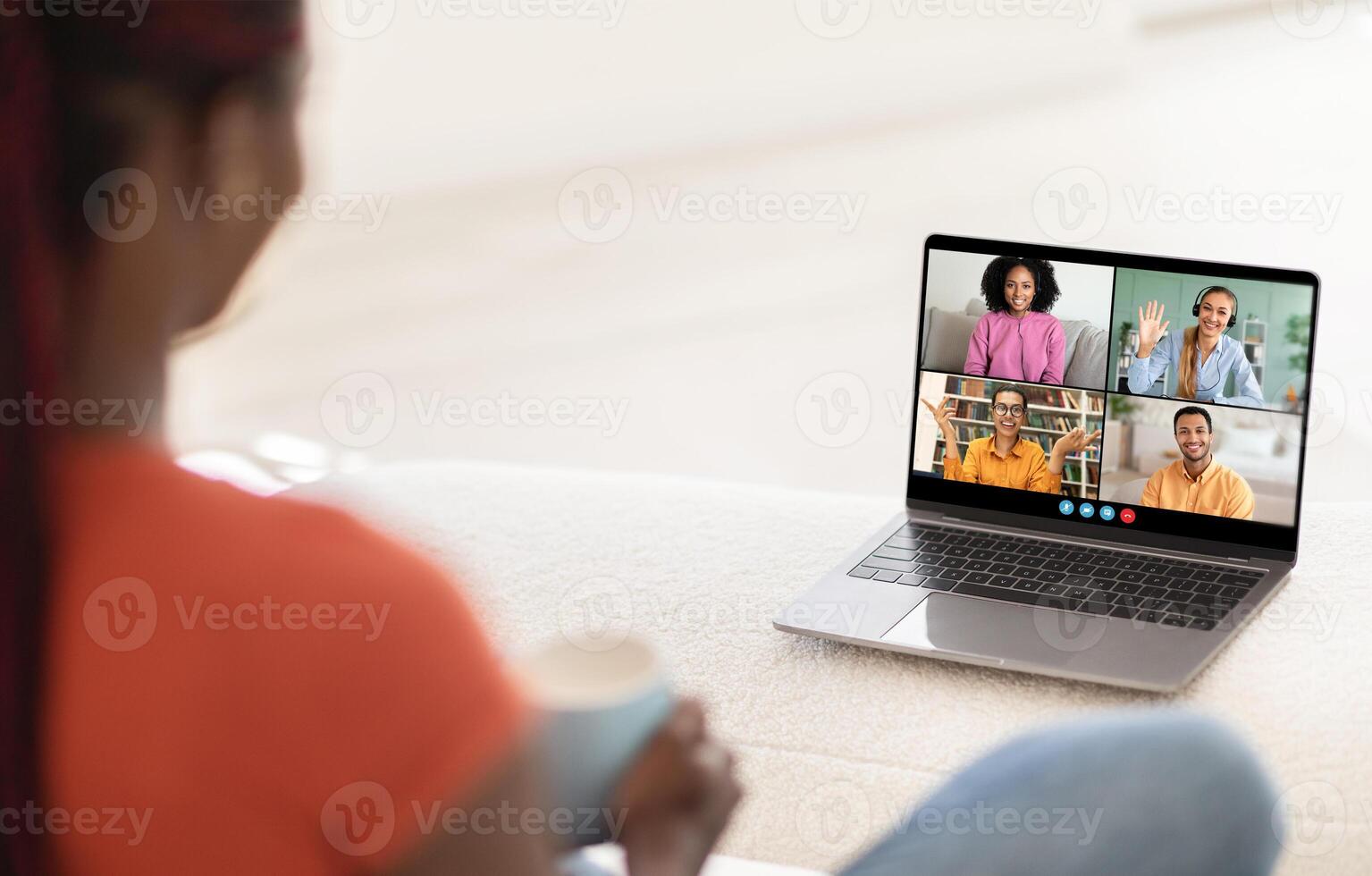 A woman sits on a couch with a laptop in front of her, attending a virtual meeting with four other people on the screen. The laptop is open and displaying a call with four participants photo